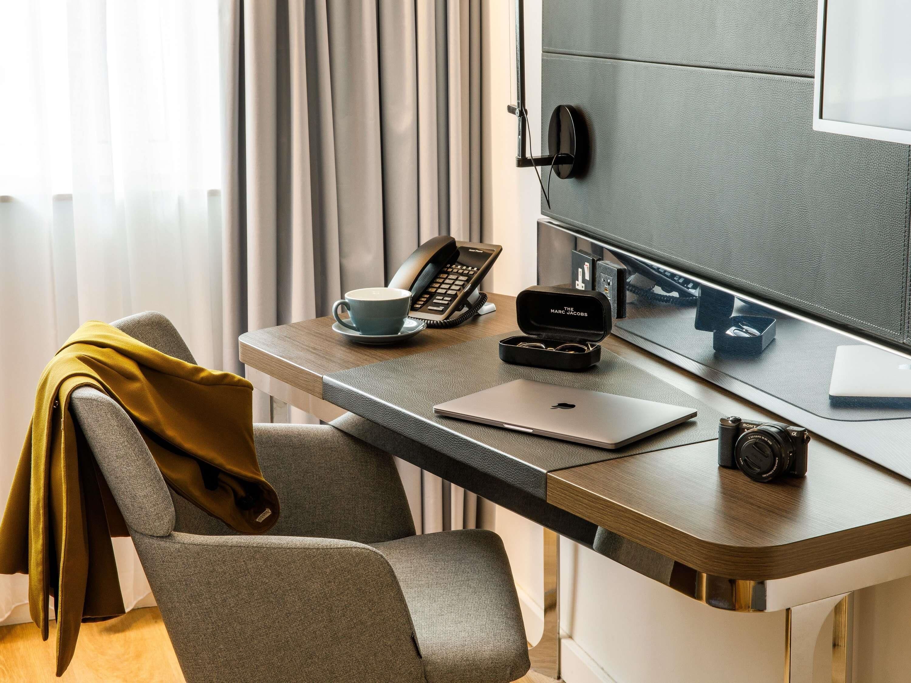 A modern hotel desk with a laptop, a camera, a phone, and a cup.
