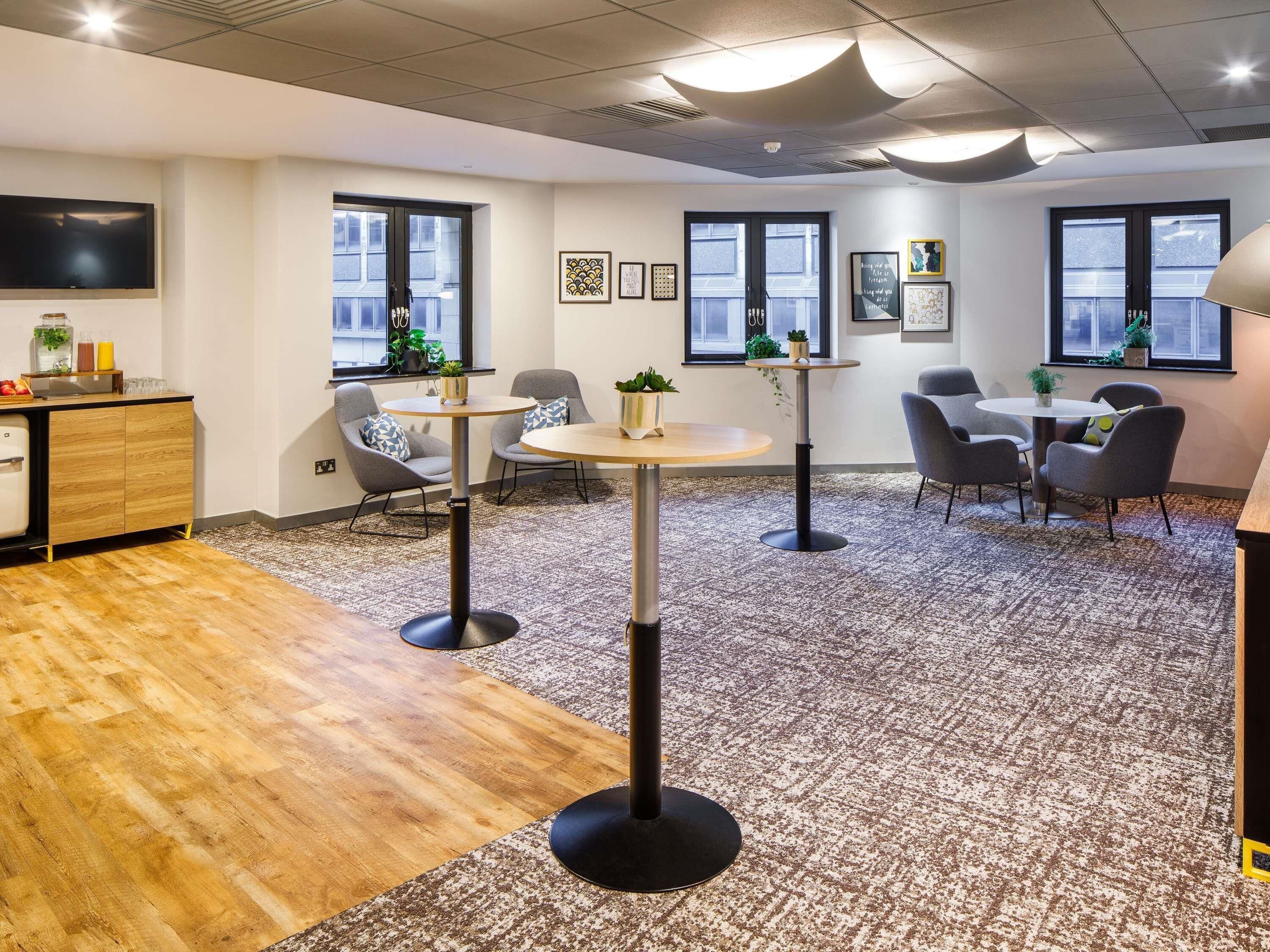 A modern office space with a wooden desk, two round tables, grey chairs, and a patterned carpet.