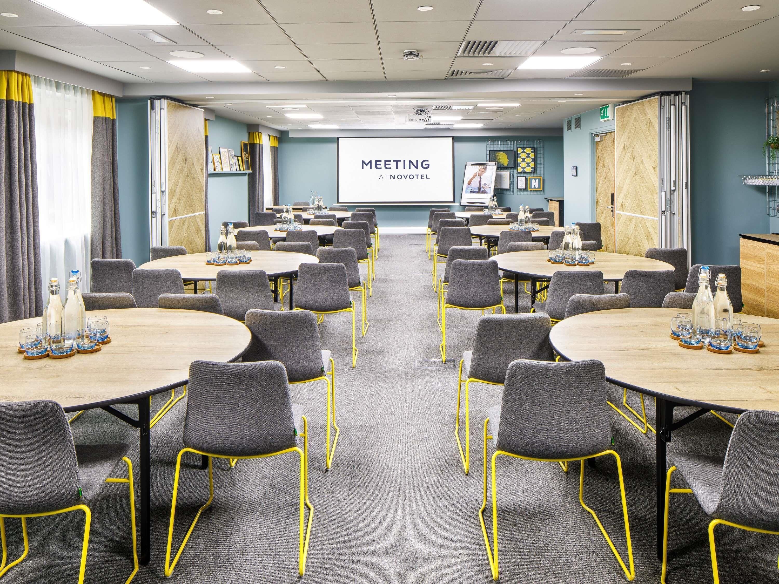 A meeting room with tables and chairs, a projector screen, and a presentation area.
