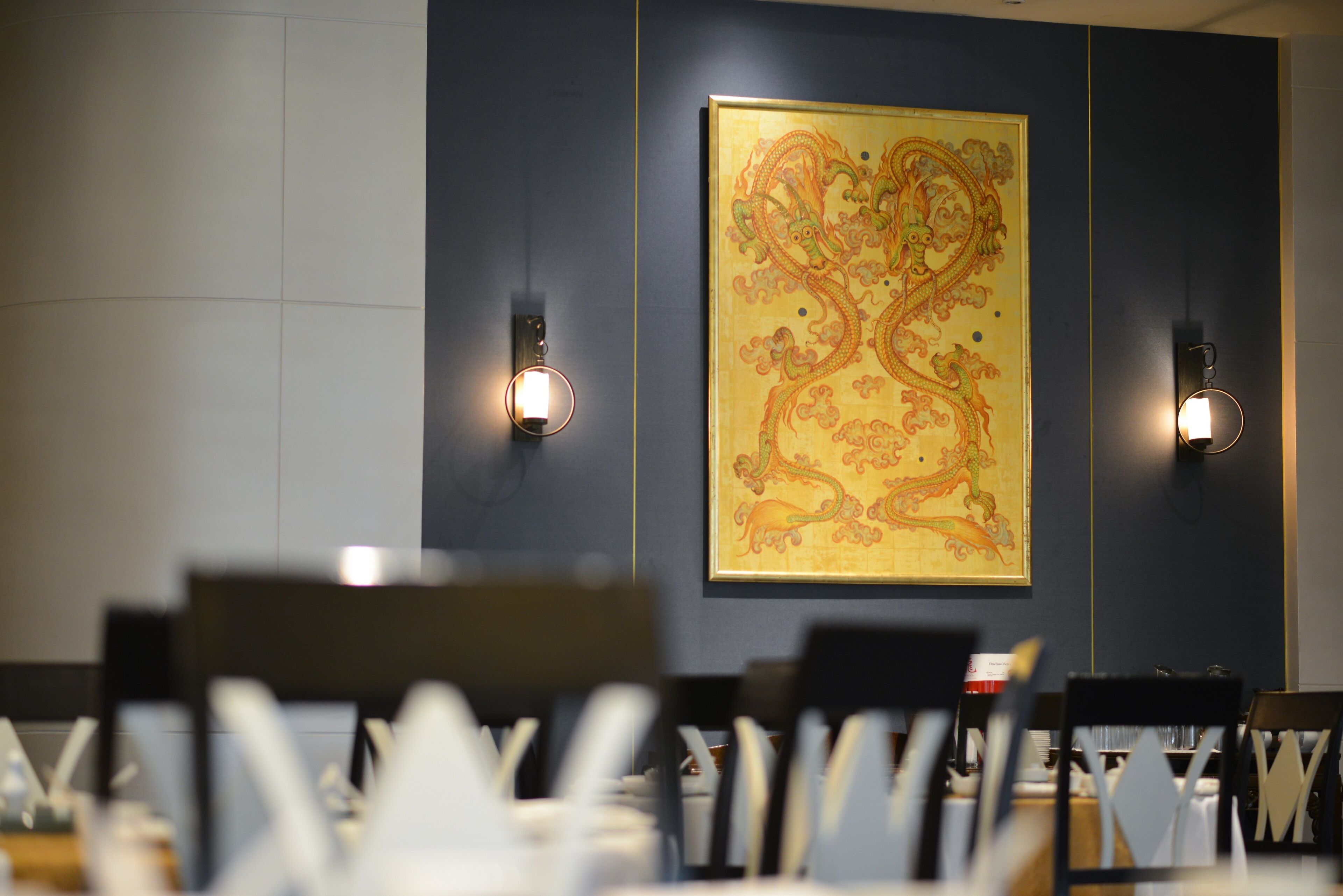A dining area with tables and chairs, a large decorative wall painting, and wall-mounted lighting fixtures.