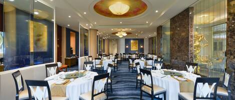 Une salle de banquet avec des tables rondes dressées pour le repas, agrémentées de nappes blanches et de chaises, et recouverte d’un tapis à motifs.
