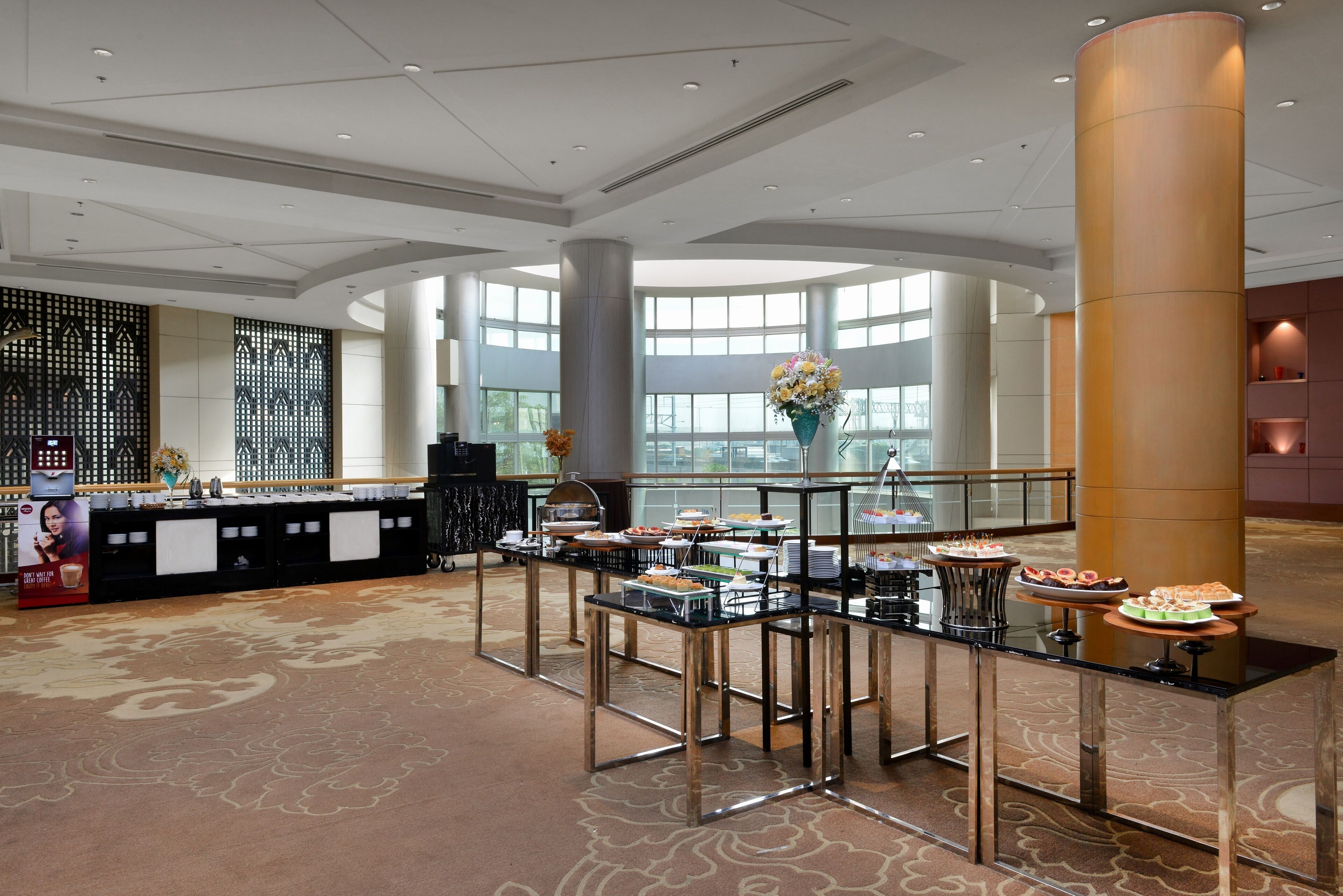 A spacious hotel lobby with a reception desk, a buffet area with various food items, large windows, and a decorative wall.