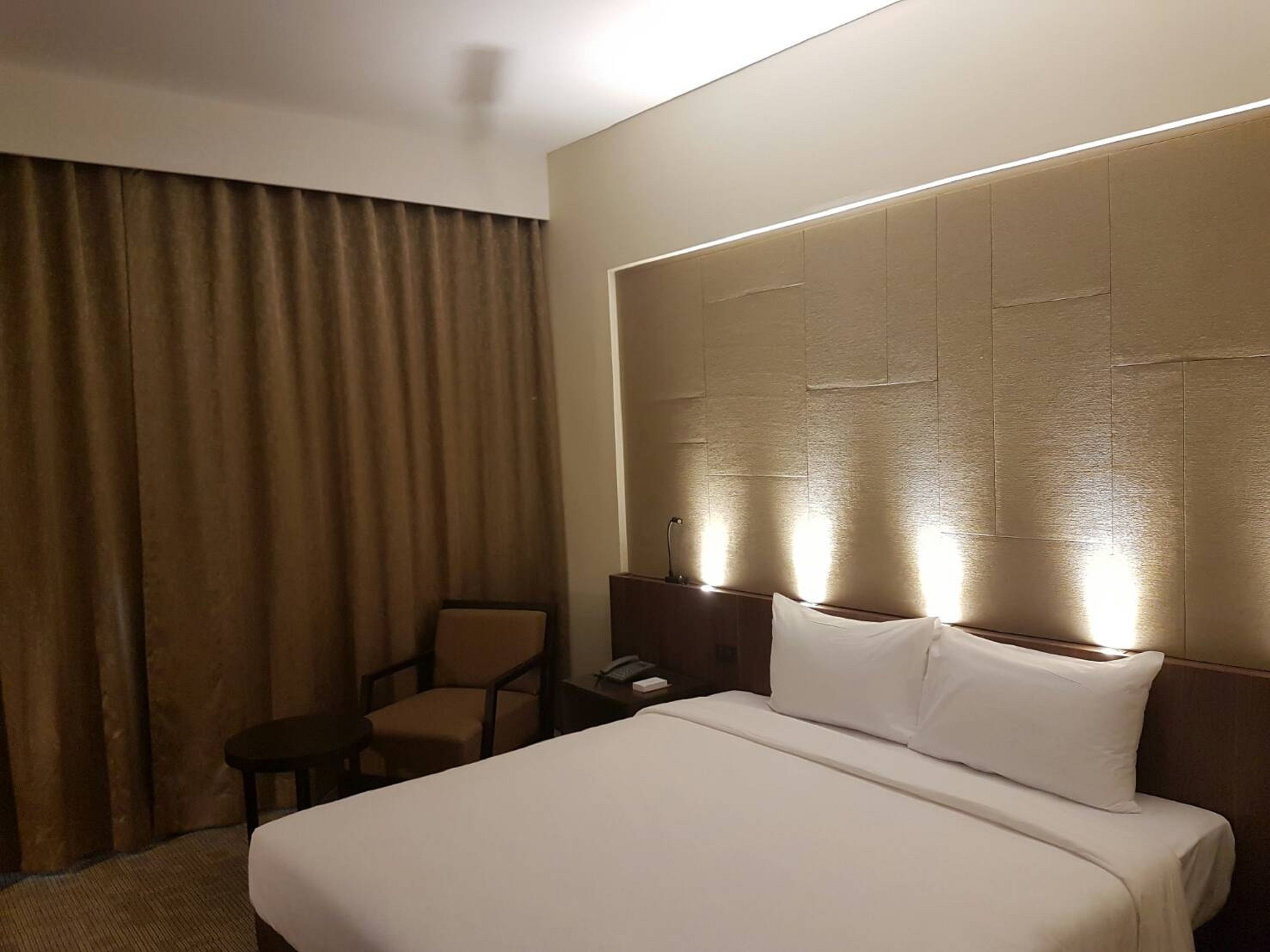 A hotel room with a large bed, a chair, a nightstand, and a headboard with built-in lighting.