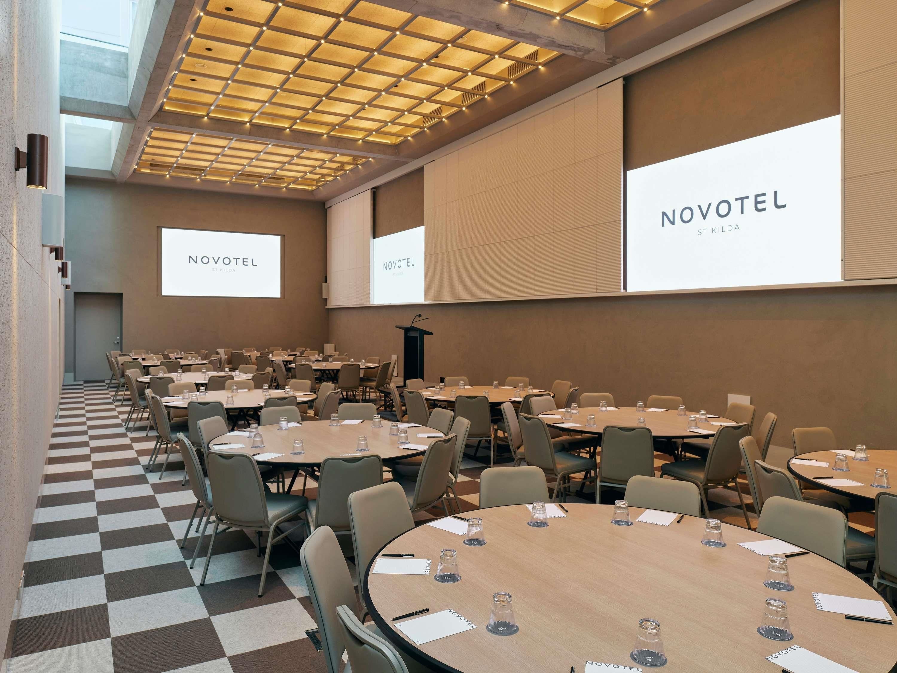 A conference room with multiple tables and chairs, a checkered floor, and large screens displaying 'NOVOTEL ST KILDA'.