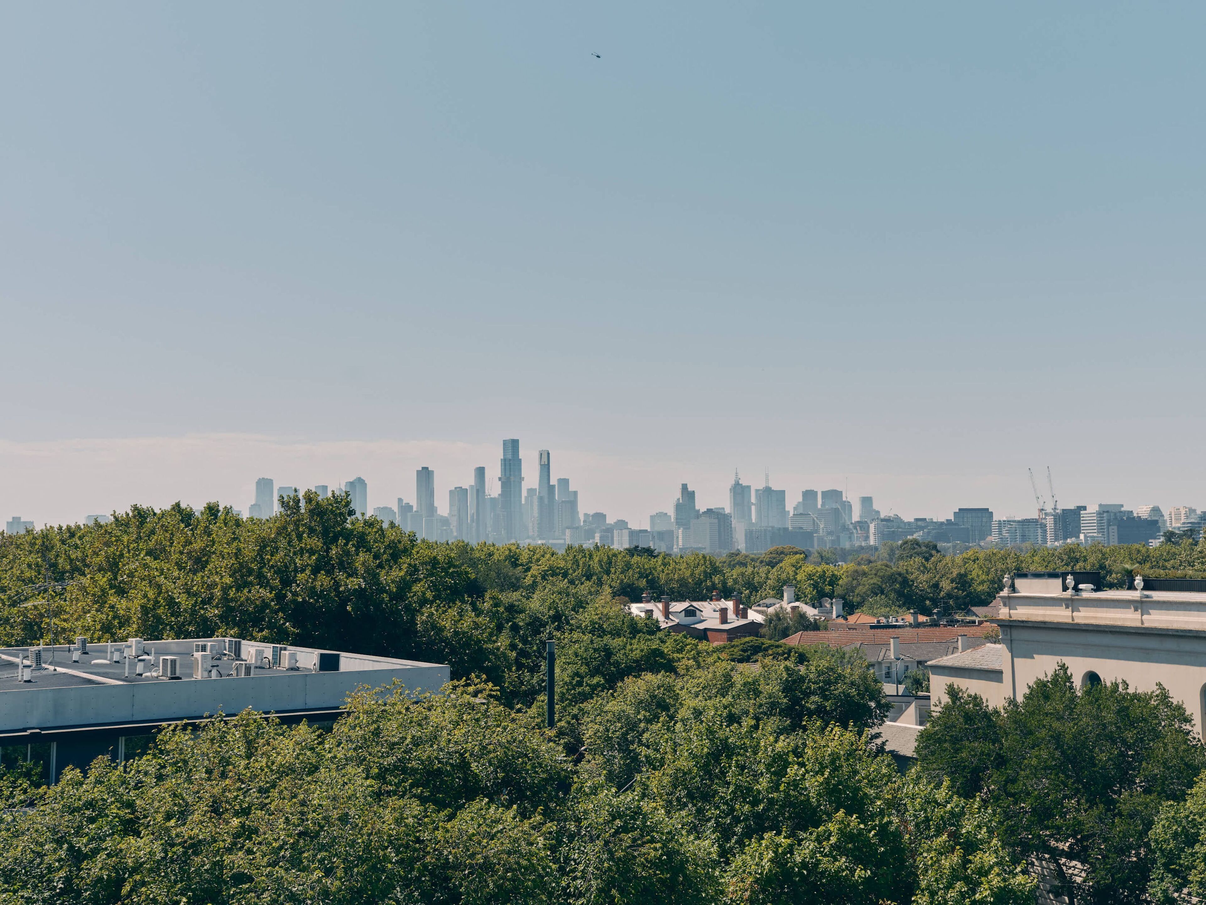 A city skyline with high-rise buildings, a rooftop with air conditioning units, and dense green trees in the foreground.