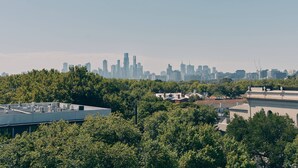 Silhueta urbana com edifícios altos, um terraço com aparelhos de ar condicionado e árvores verdes densas em primeiro plano.