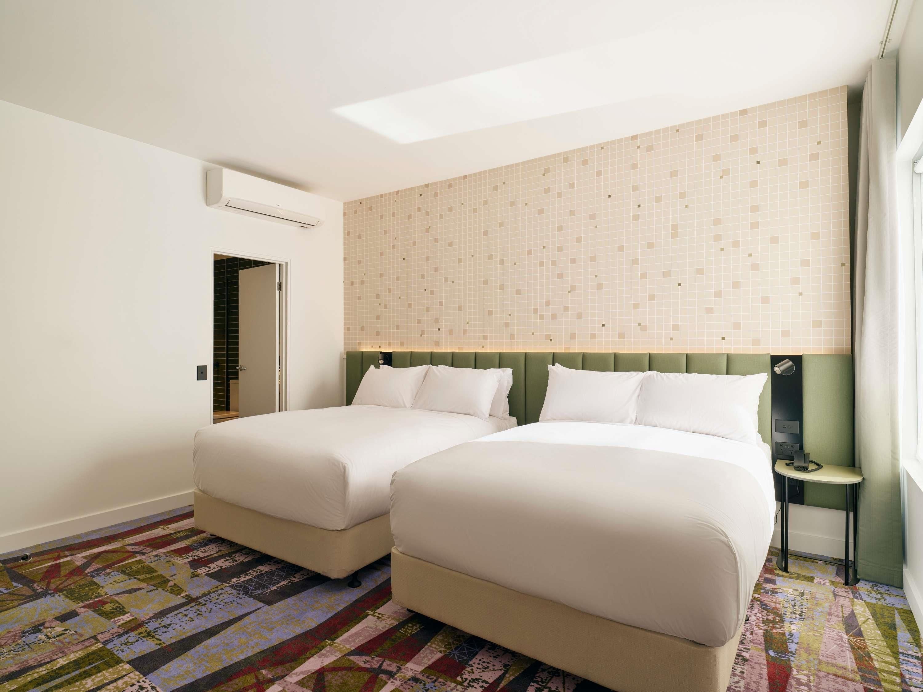 A hotel room with two beds, a patterned carpet, a green headboard, and a window with curtains.