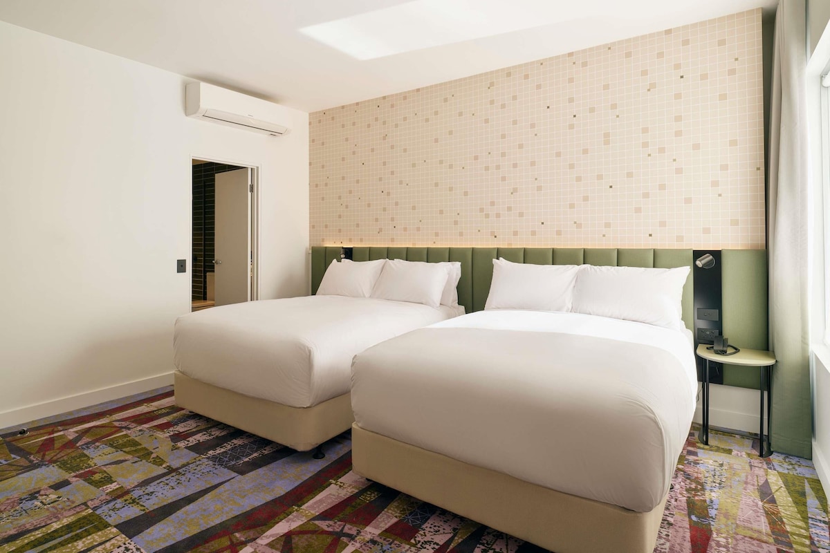 A hotel room with two beds, a patterned carpet, a green headboard, and a window with curtains.