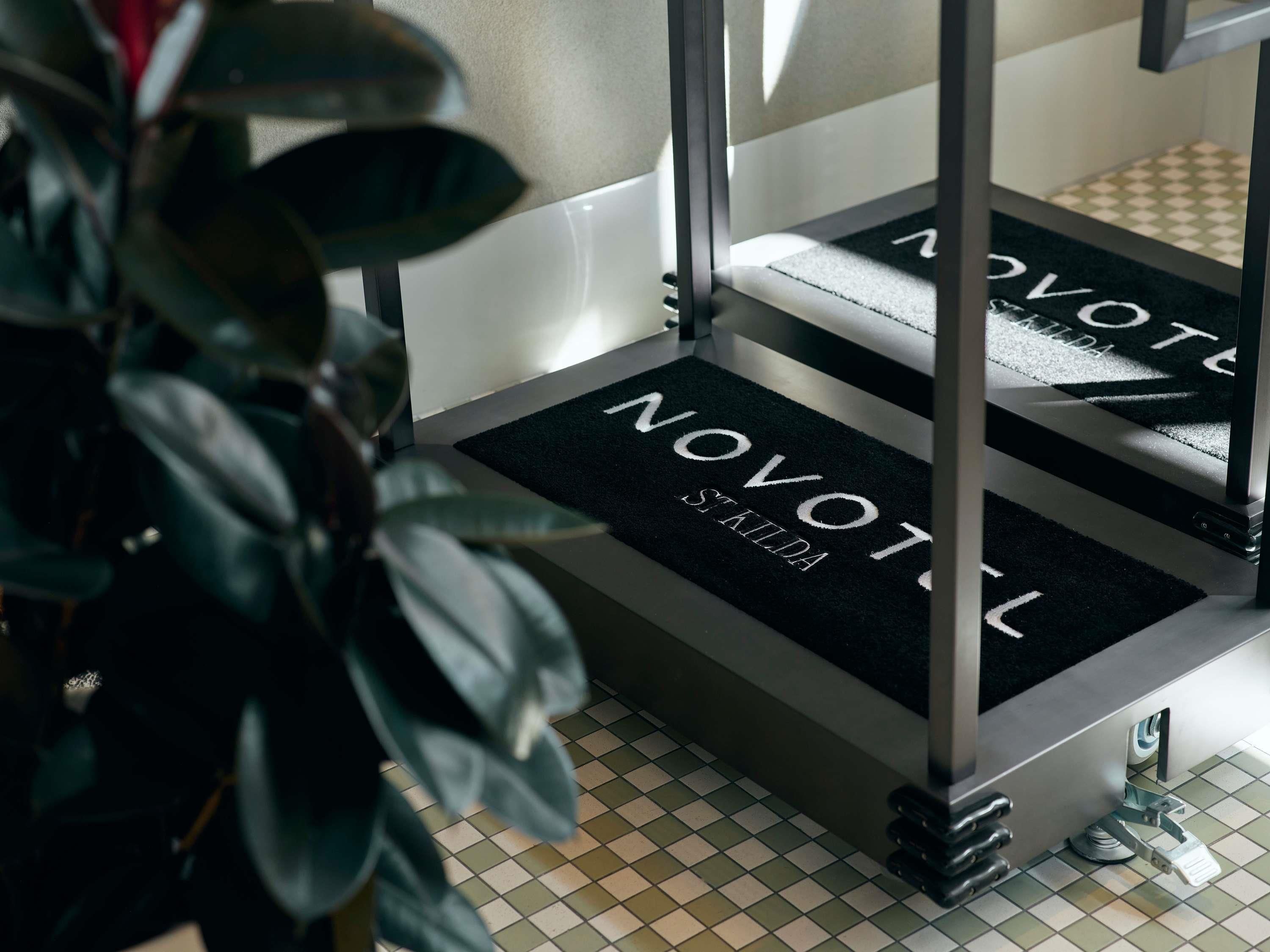 Una escalera con un tapete negro que muestra los textos 'NOVOTEL' y 'ST. KILDA', que lleva a una habitación con piso de baldosas.