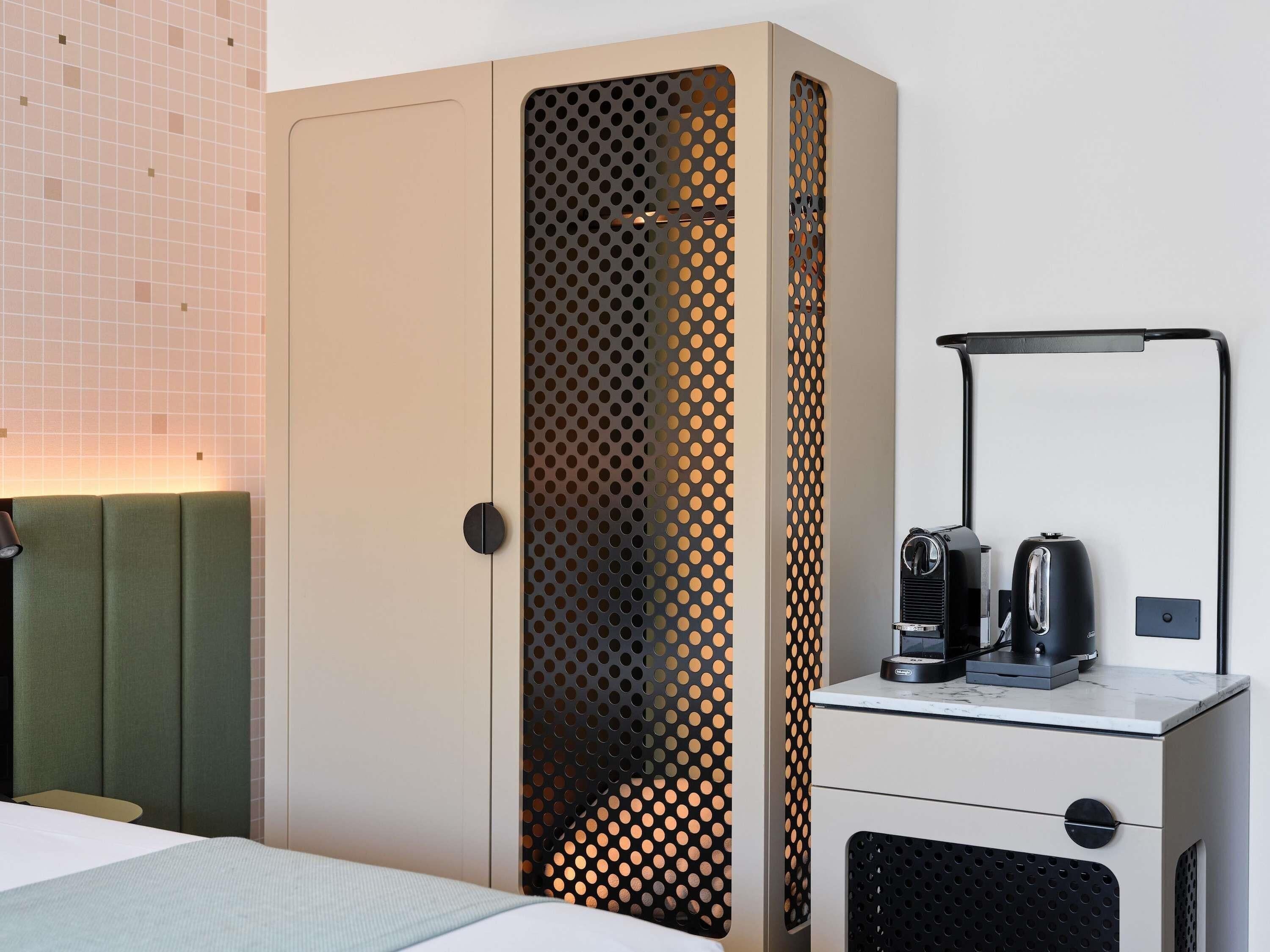 A hotel room with a bed, a green upholstered headboard, a wardrobe with a perforated design, a desk with a coffee maker, and a mirror.