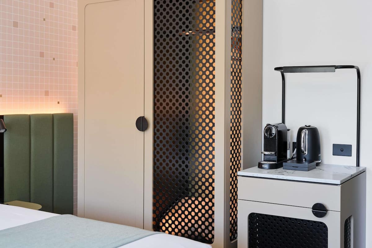 A hotel room with a bed, a green upholstered headboard, a wardrobe with a perforated design, a desk with a coffee maker, and a mirror.