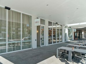 Uma área moderna de estar ao ar livre, com mesas e cadeiras, grandes janelas e uma placa indicando um restaurante.