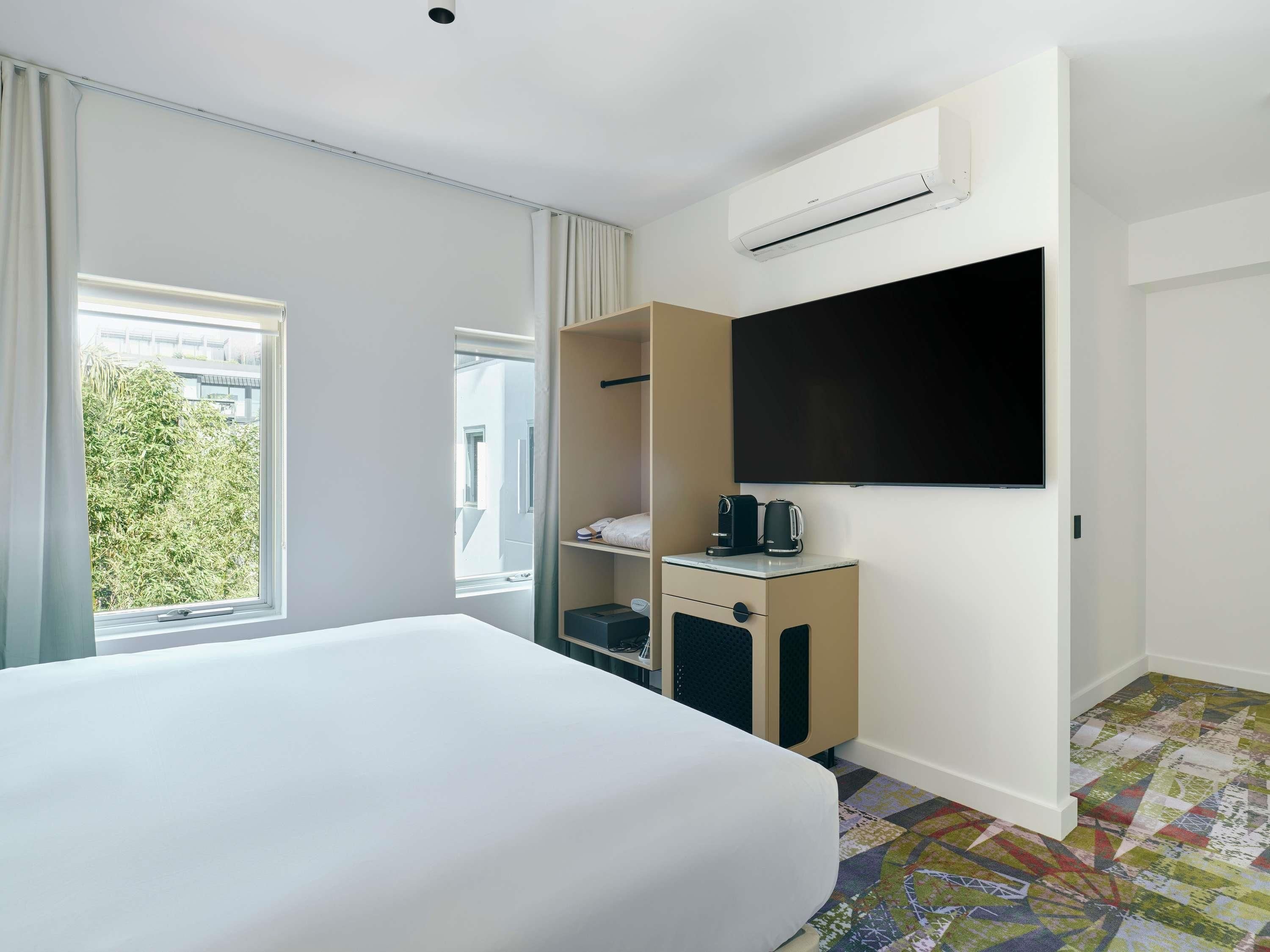 A hotel room with a bed, a wardrobe, a television, and a window with a view of greenery.