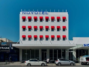 Hotel com fachada moderna, várias janelas com toldos vermelhos e céu azul límpido.
