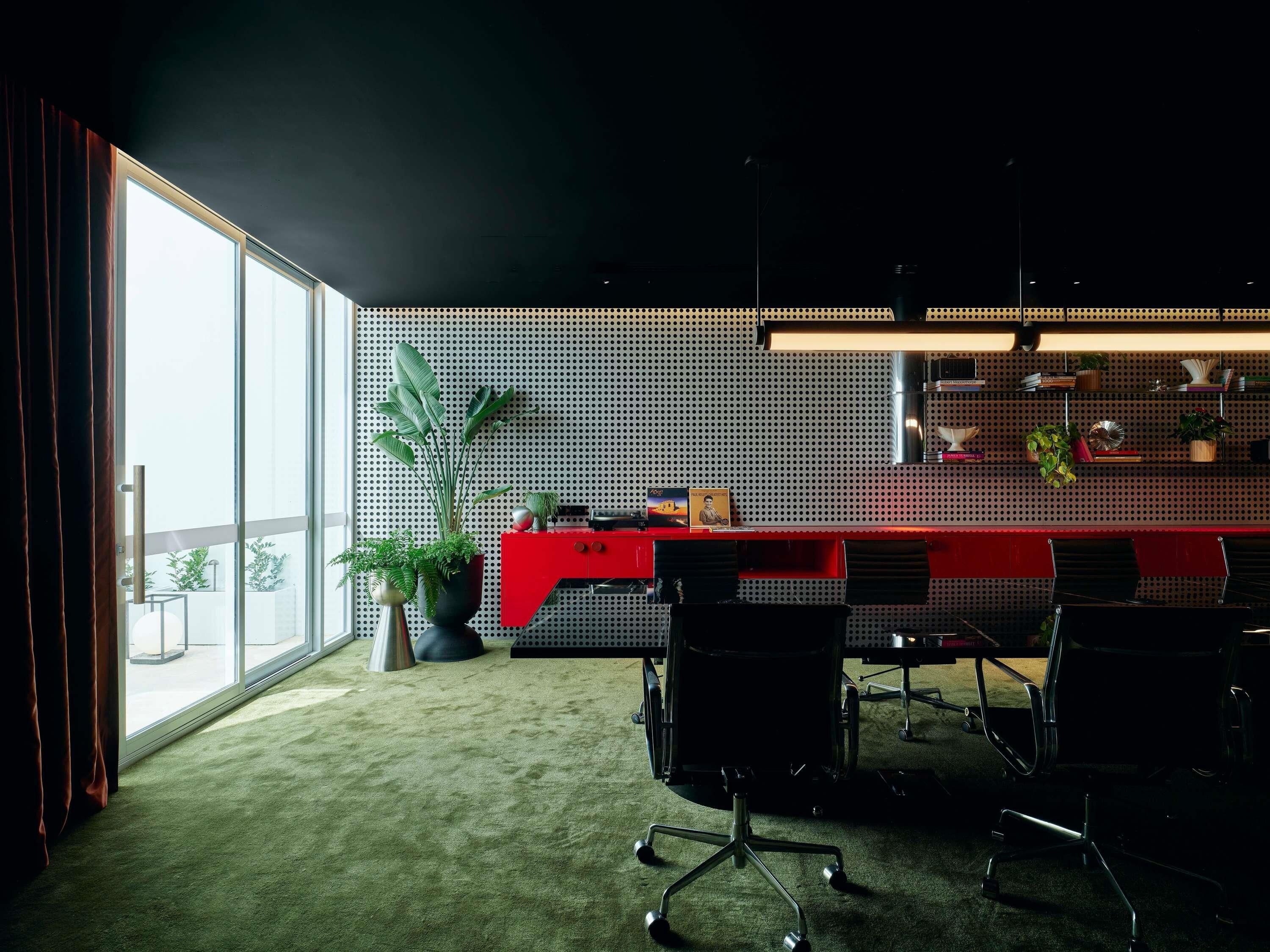 A modern office space with a red desk, black chairs, a large potted plant, and a wall with a perforated pattern.