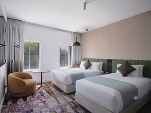 A hotel room with two beds, a chair, a small table, and a window with curtains. - Novotel St Kilda (St Kilda)