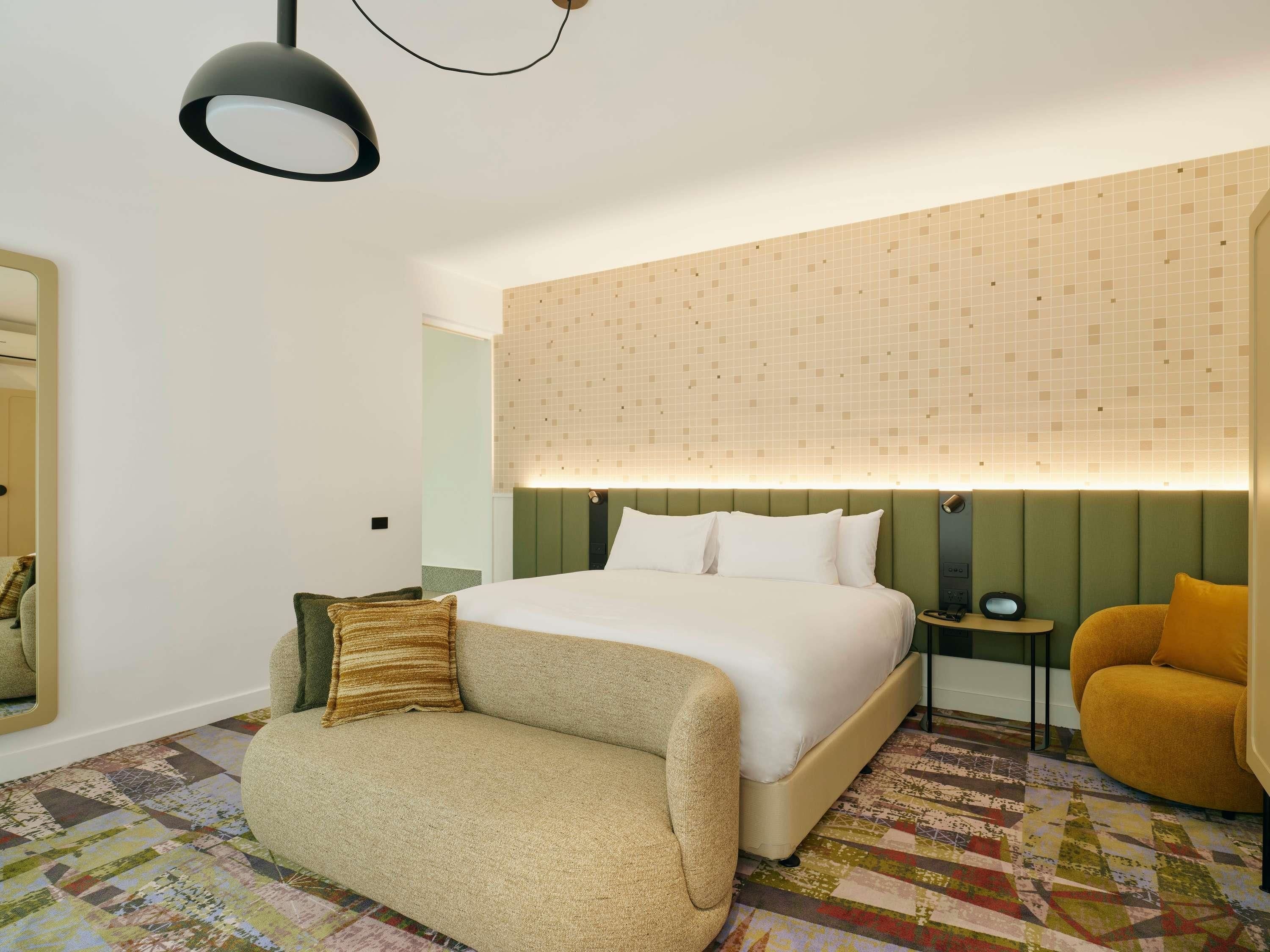 A hotel room with a bed, a sofa, a chair, a nightstand, and a mirror.