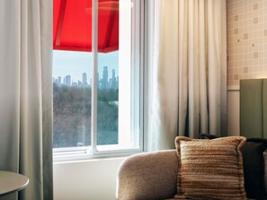 A room with a window, a view of a city skyline, a chair, and a small table. - Novotel St Kilda (St Kilda)