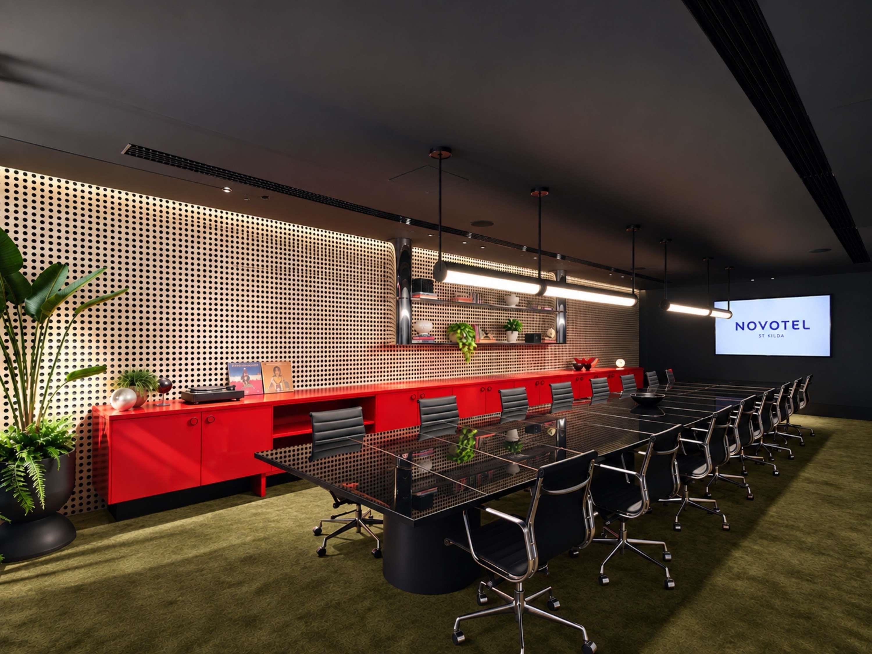 A modern conference room with a long table, red accents, and a projector screen displaying 'NOVOTEL ST KILDA'.