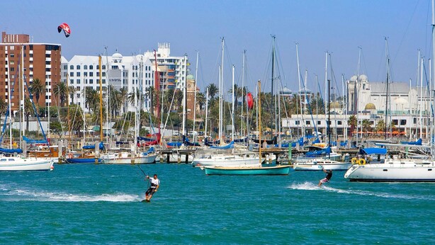 Uma marina com vários barcos atracados, uma pessoa praticando wakeboard e edifícios ao fundo.