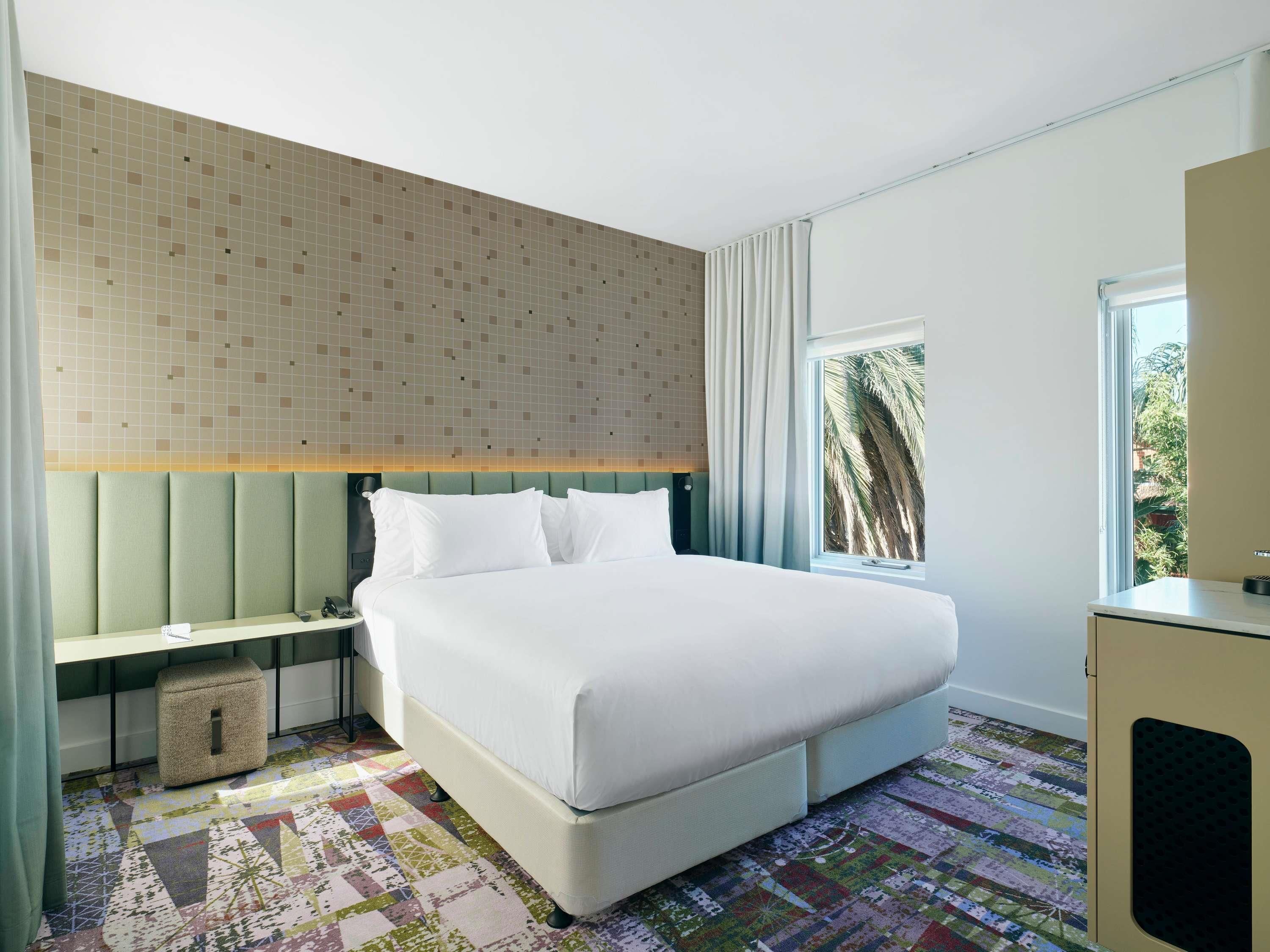 A hotel room with a large bed, a nightstand, a small stool, a window with a view of palm trees, and a patterned floor.