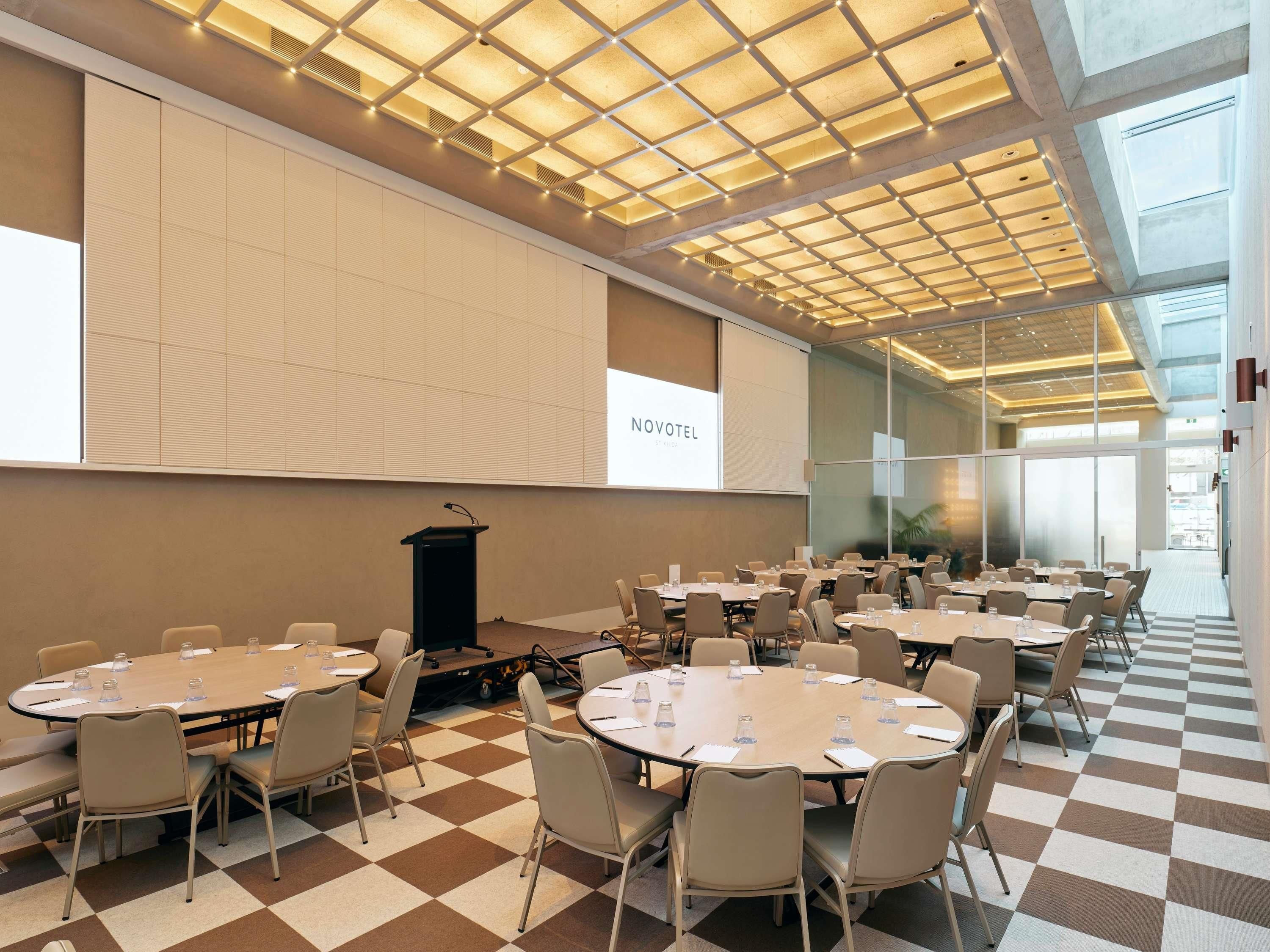 A conference room with multiple tables and chairs, a podium, and a large screen displaying 'NOVOTEL ST. LUCIA'.