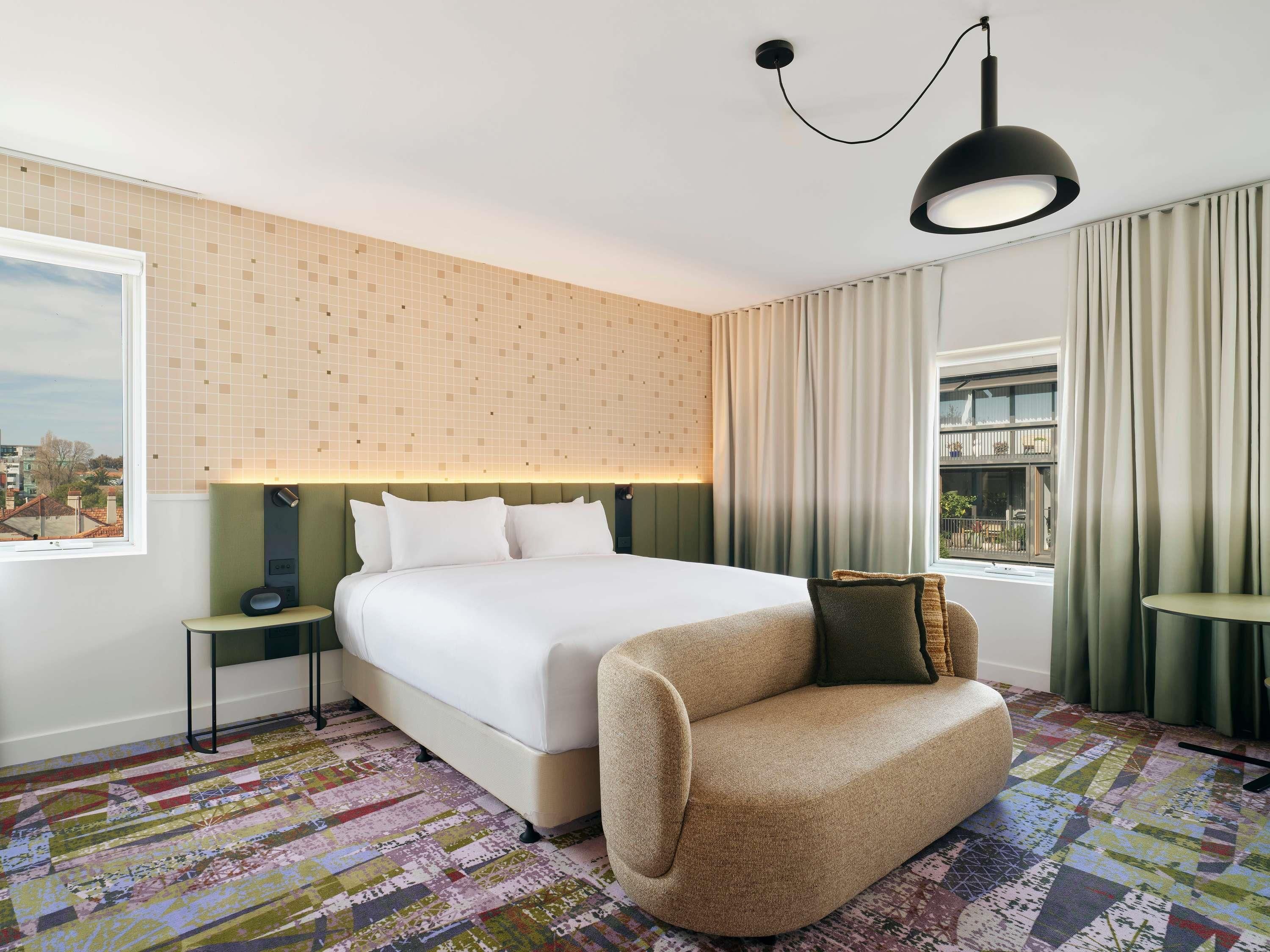 A hotel room with a large bed, a green headboard, a patterned rug, a sofa, and a view of the city through the window.