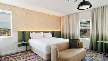 A hotel room with a large bed, a green headboard, a patterned rug, a sofa, and a view of the city through the window.