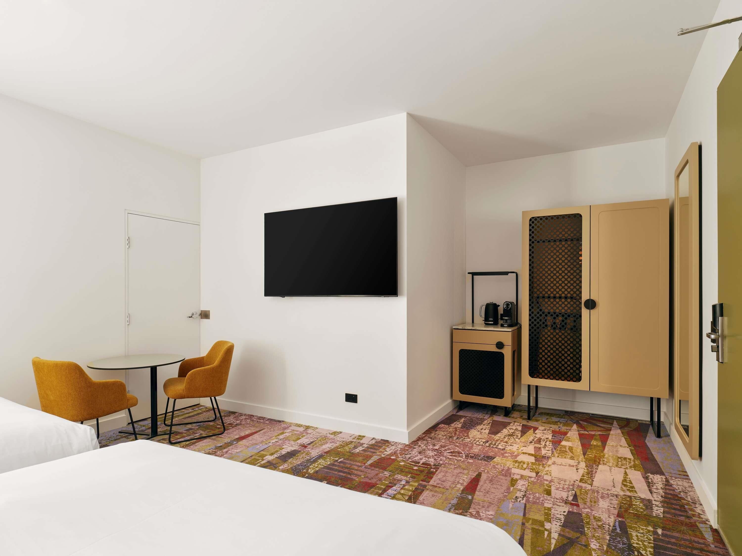 A hotel room with a bed, a desk with two chairs, a television mounted on the wall, a wardrobe, and a patterned rug.