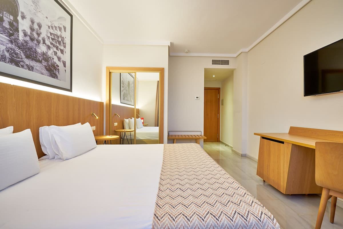 Quarto de hotel com cama, mesas de cabeceira, escrivaninha, cadeira, televisão e um quadro na parede.
