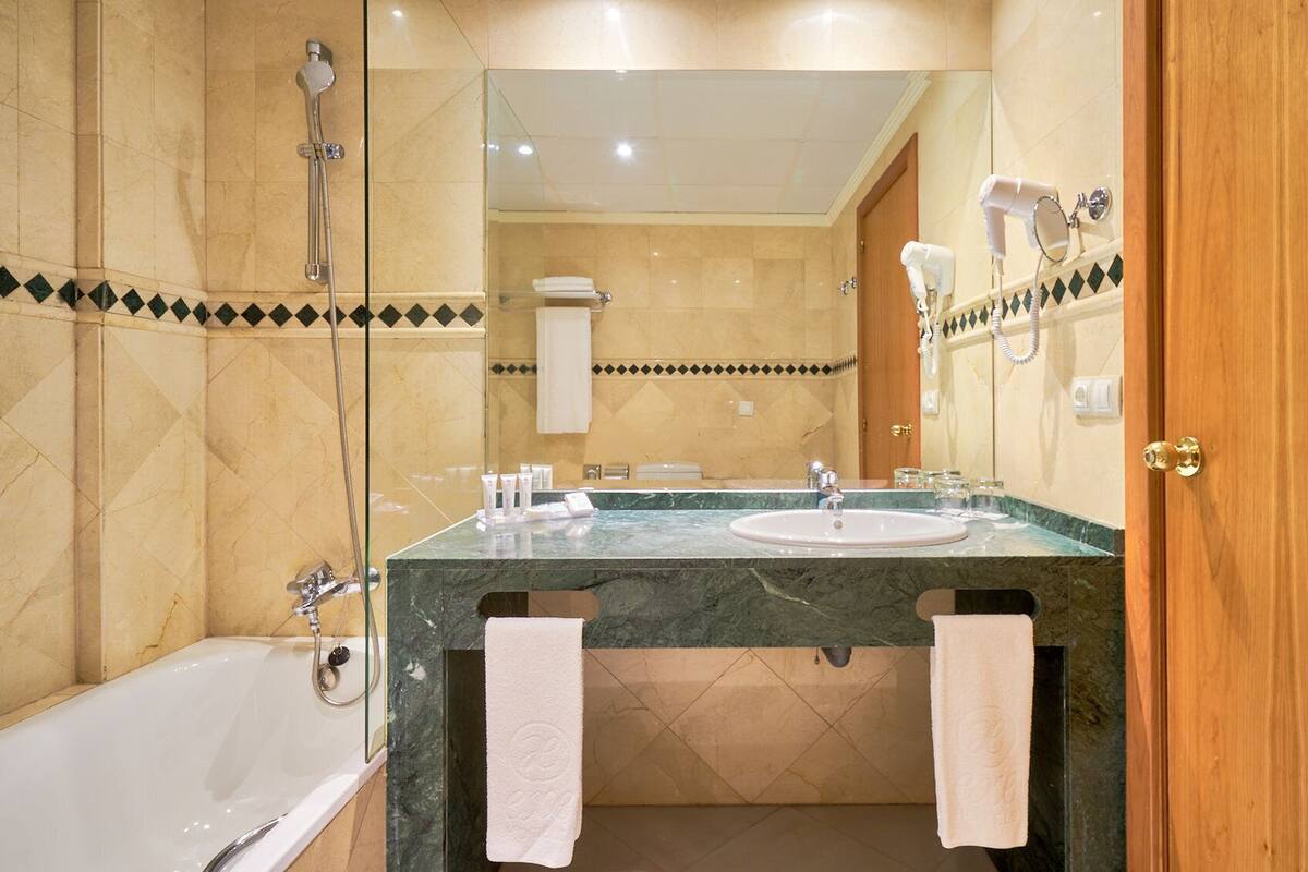 Casa de banho com banheira, lavatório e chuveiro. Toalhas penduradas na parede e um secador de cabelo no balcão.
