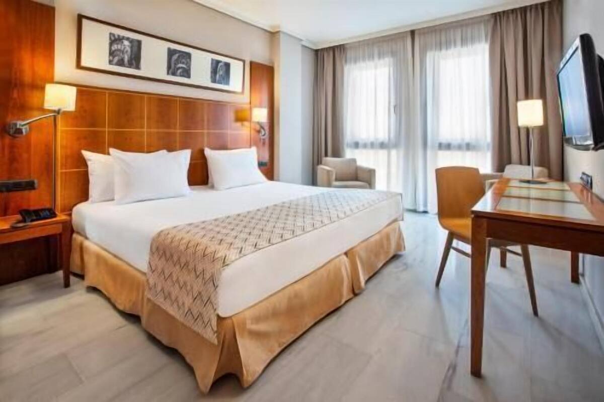 Quarto de hotel com uma cama grande, uma escrivaninha, uma cadeira, uma televisão e uma janela com cortinas.