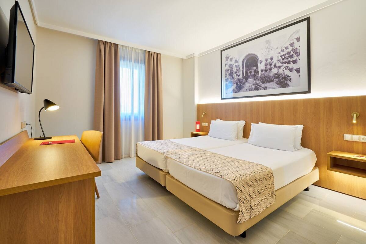 Quarto de hotel com cama, mesas de cabeceira, escrivaninha, cadeira, televisão e um quadro na parede.