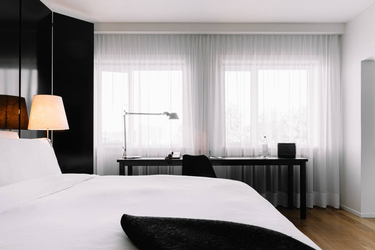 Una habitación de hotel moderna con una cama grande, un escritorio y una ventana con cortinas translúcidas.