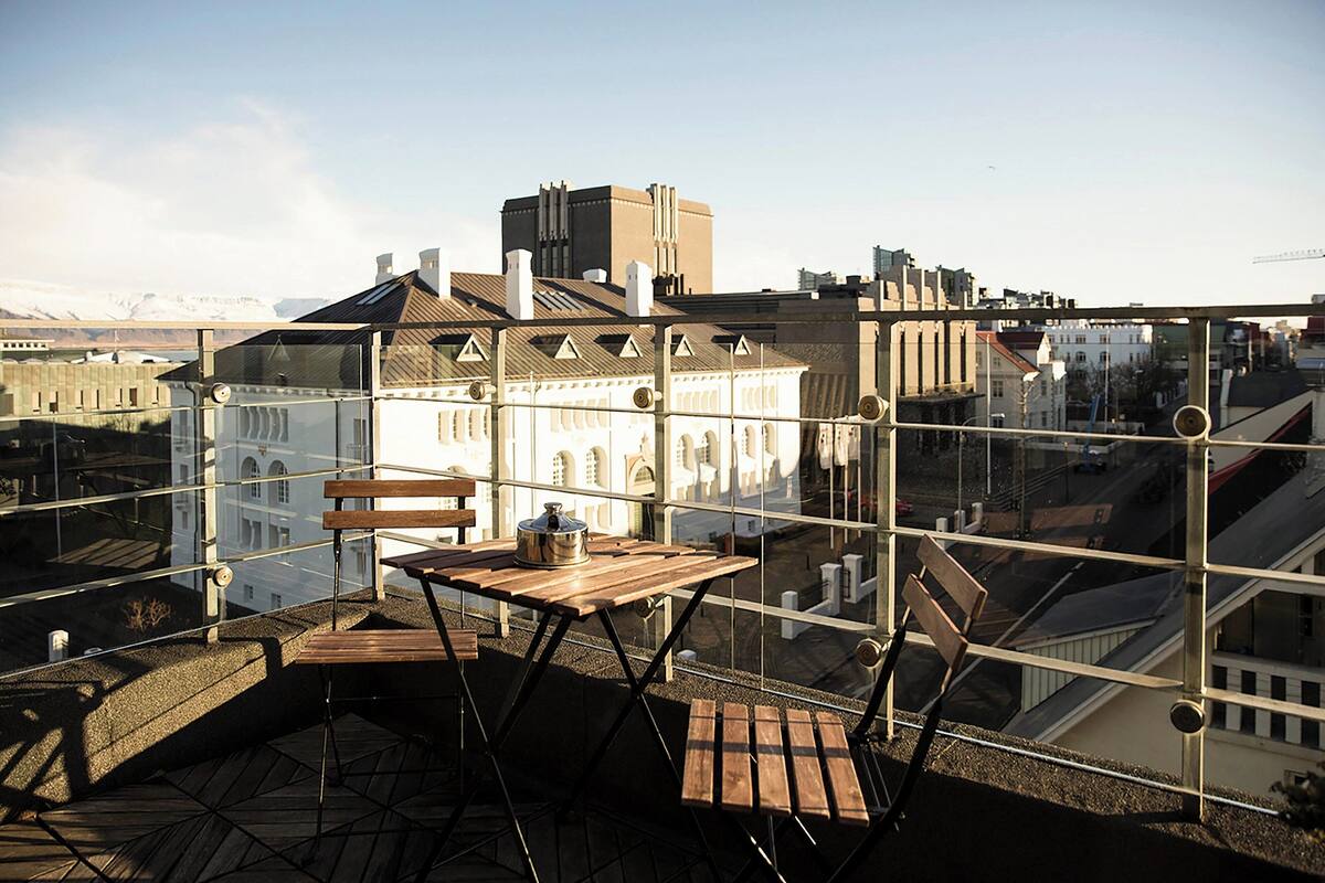 Una terraza en la azotea con mesa y sillas de madera, una olla de metal y vistas a edificios de la ciudad.
