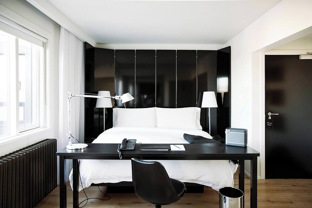 Una habitación de hotel moderna con cama, escritorio, dos lámparas, una silla, un radiador y una puerta.