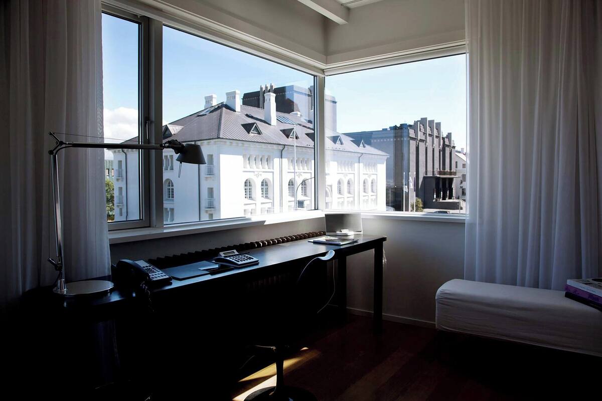 Una habitación con escritorio, teclado, lámpara y vistas a edificios a través de amplios ventanales.