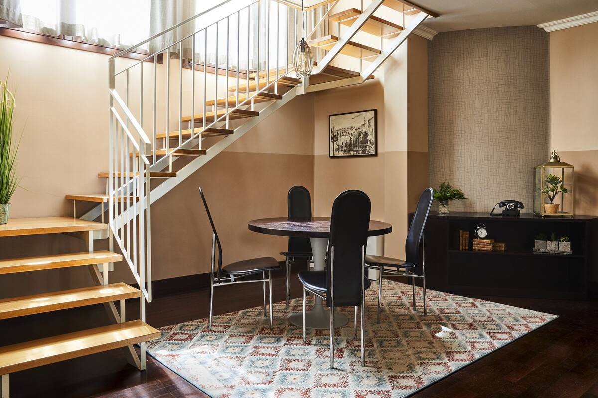 Eine Treppe mit Holzhäusern und weißen Geländern, ein runder Esstisch mit schwarzen Stühlen und ein gemustertes Teppich im Raum.