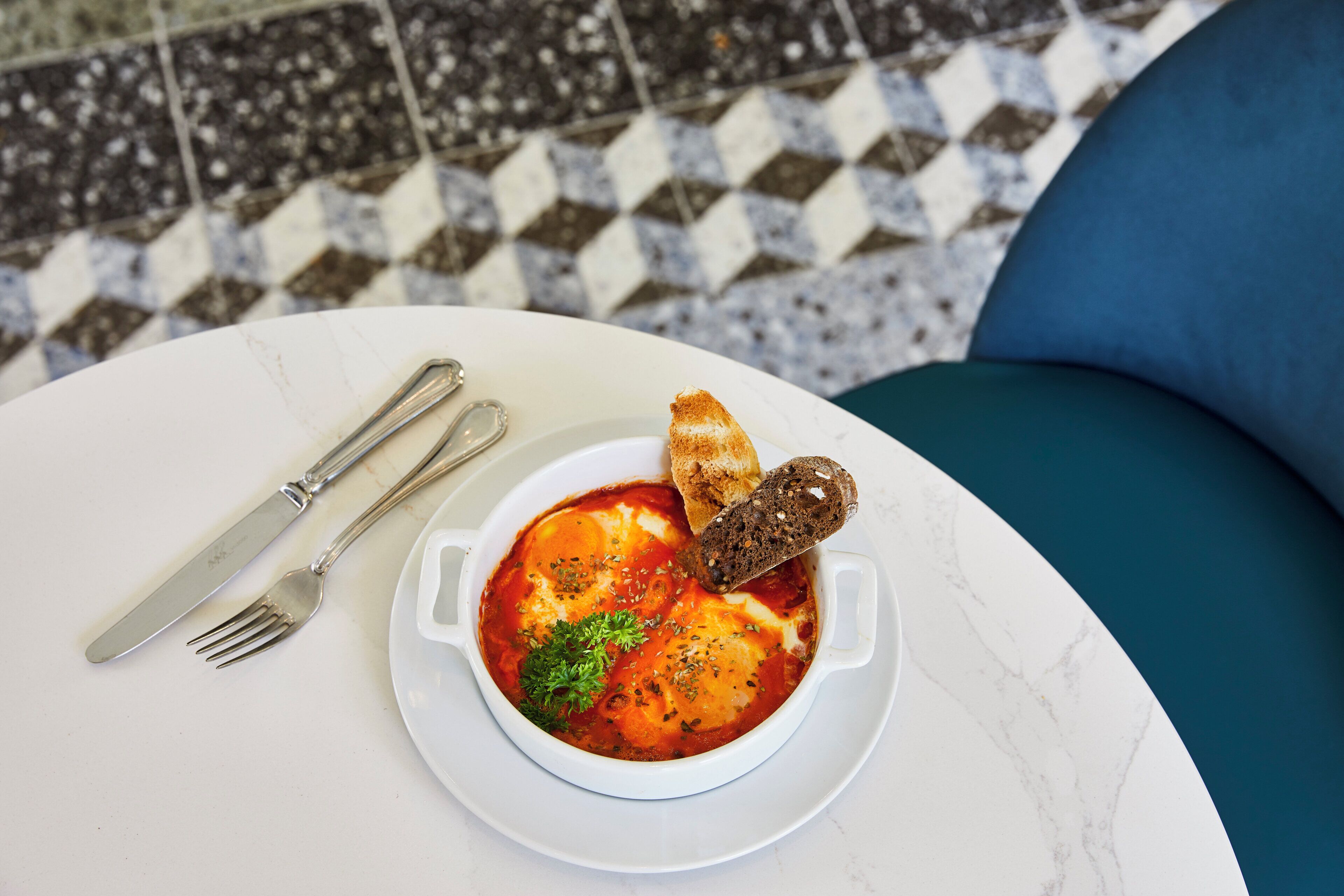 Un tazón de cerámica blanca con salsa roja, adornado con hierbas, sobre una mesa blanca con un cojín azul.