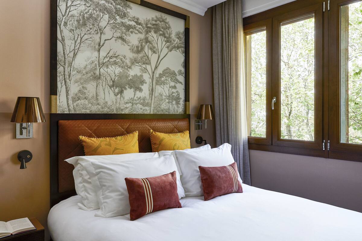 Une chambre d’hôtel avec un grand lit, une tête de lit ornée d’une fresque représentant un arbre, deux lampes de chevet et une fenêtre avec des rideaux.