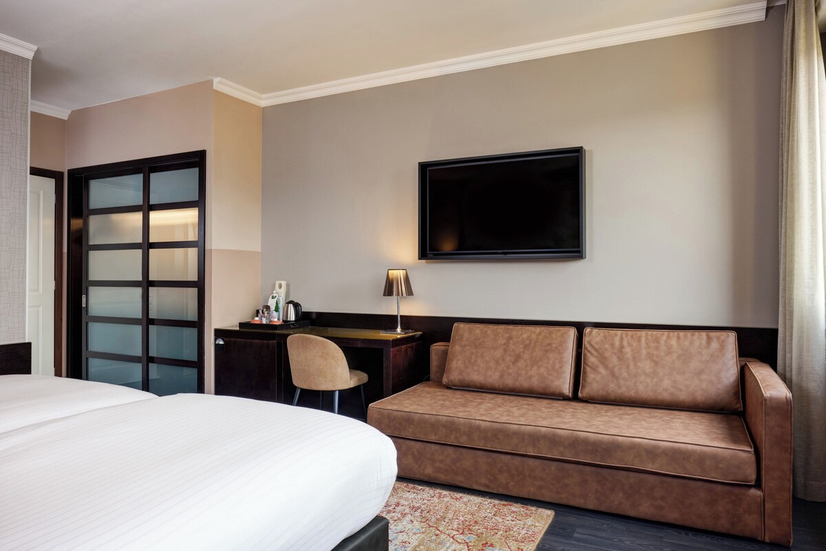 Une chambre d’hôtel comprenant un lit, un canapé, un bureau, une chaise, un téléviseur à écran plat et une porte dotée de panneaux en verre dépoli.