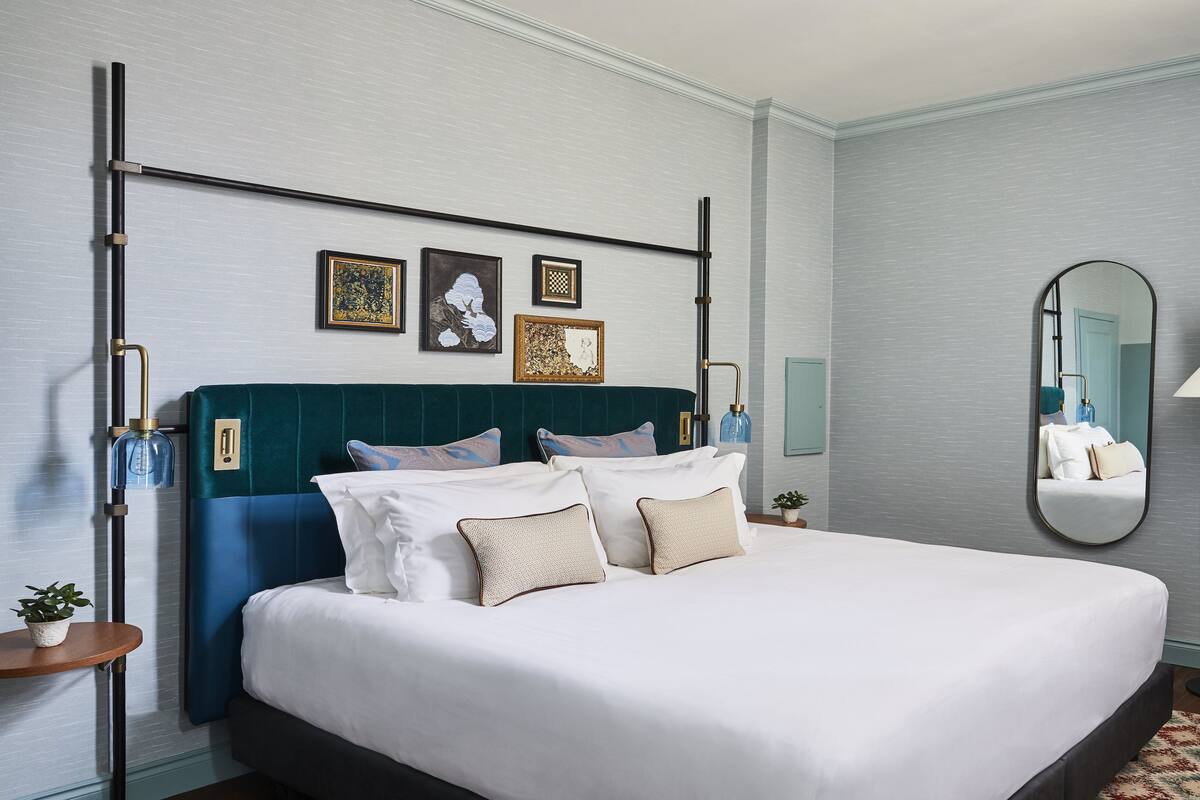 Un lit bien fait, avec du linge de lit blanc et plusieurs oreillers, une tête de lit recouverte d’un tissu turquoise et une table de chevet agrémentée d’une petite plante.