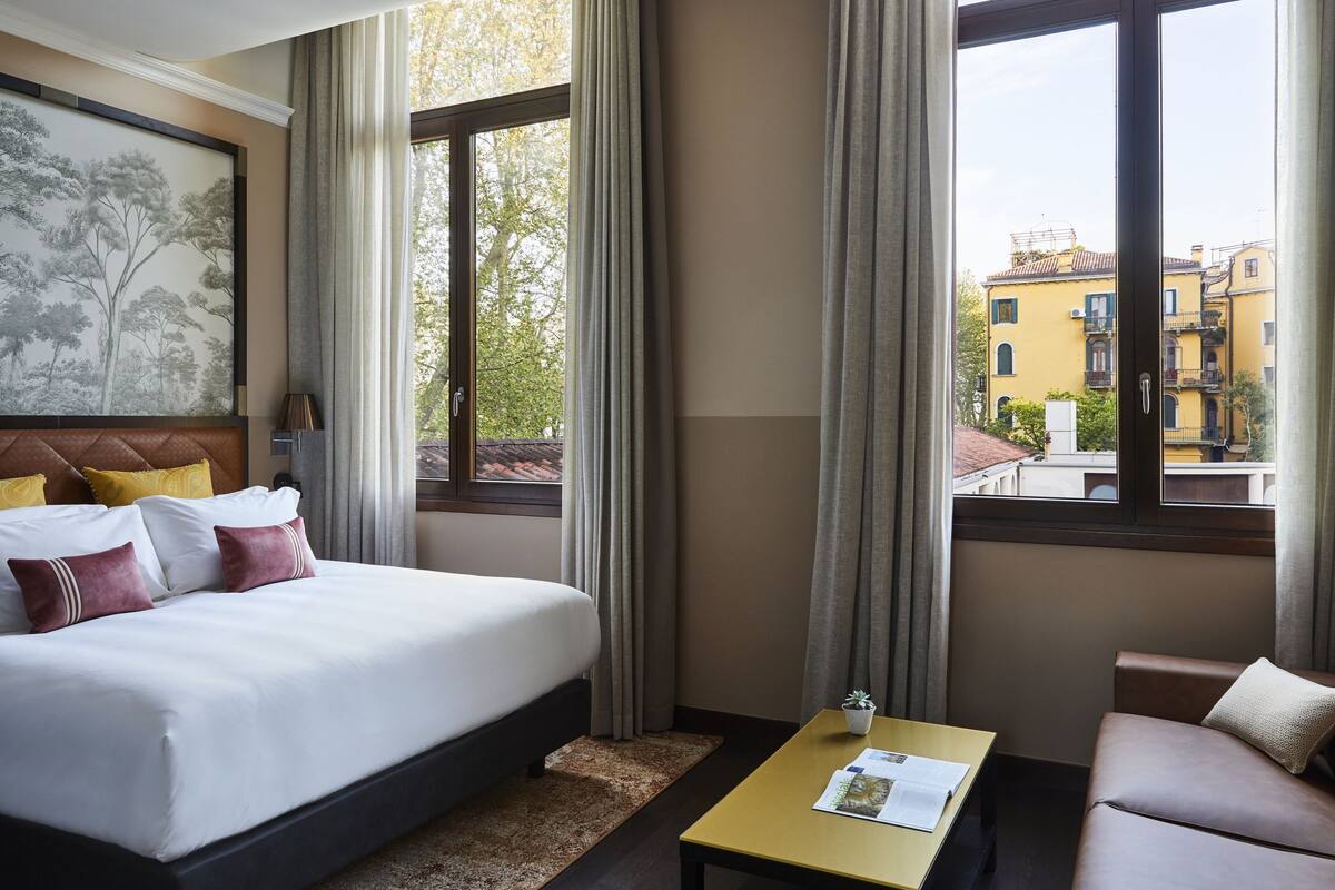 Ein Hotelzimmer mit einem großen Bett, einer kleinen Couch, einem Tisch und einem Fenster mit Blick auf Gebäude.