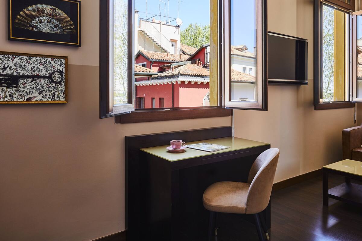 Ein Zimmer mit Schreibtisch, Sessel und einer Couch. An den Wänden hängen gerahmte Kunstwerke und über dem Schreibtisch ist ein Fernseher angebracht.