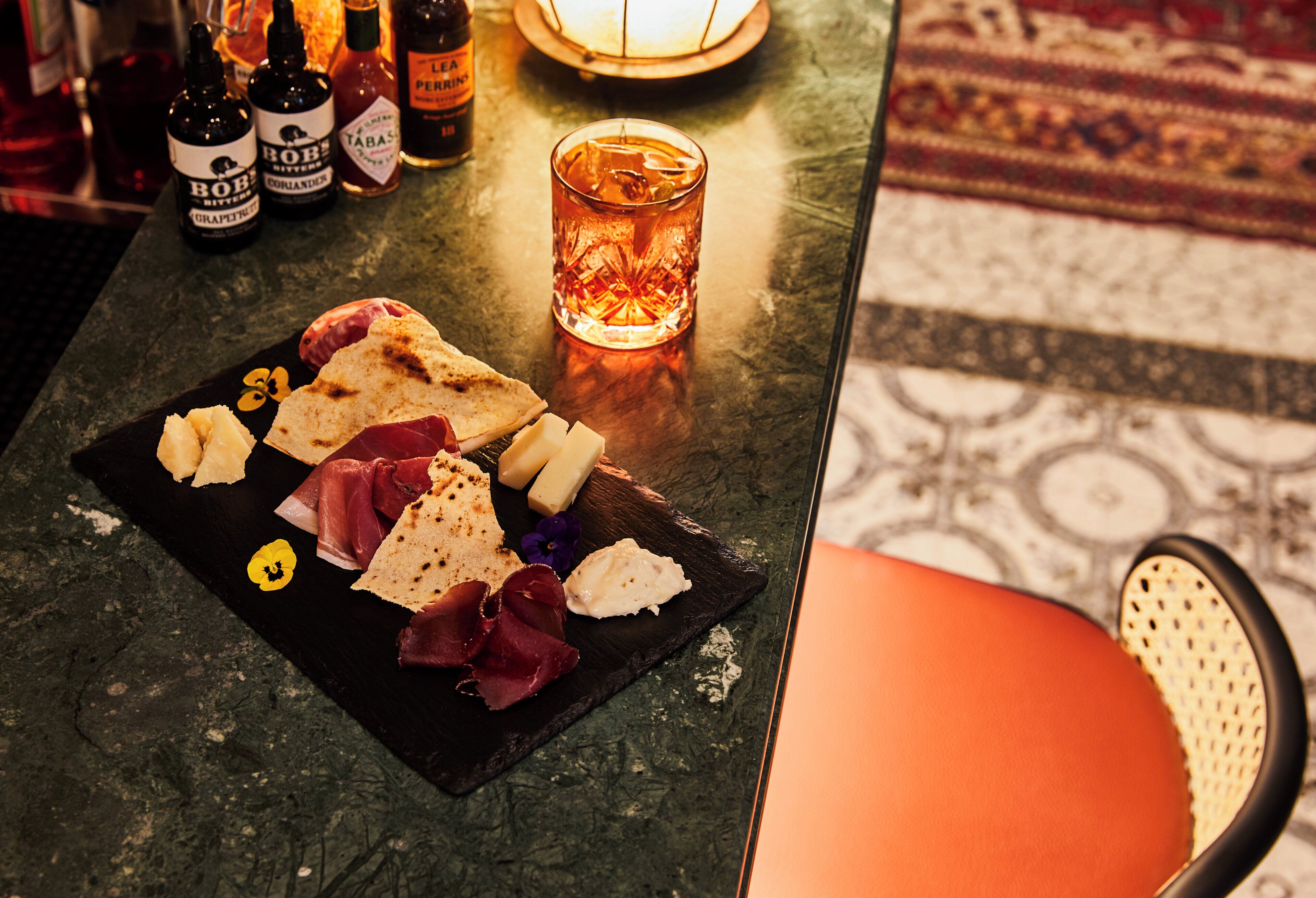 A bar area with a selection of spirits, a glass of amber-colored drink, and a charcuterie board with various meats and cheeses.