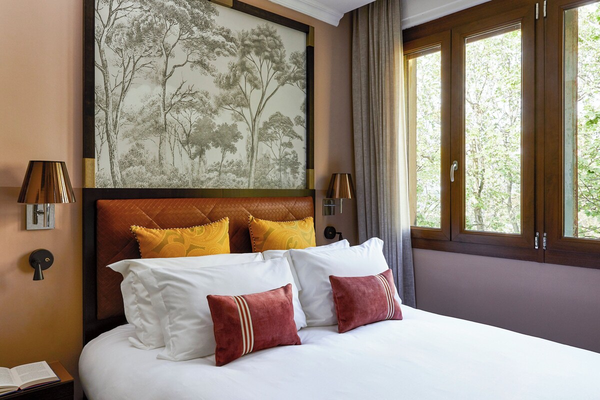 Une chambre d’hôtel avec un grand lit, une tête de lit ornée d’un motif, une œuvre d’art accrochée au mur et une fenêtre avec des rideaux.
