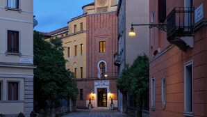 Eine schmale Straße mit Häusern an beiden Seiten, darunter ein markantes Gebäude aus rotem Backstein mit einem Uhrturm und einem Schild, das "Calle Della Carmine" zeigt.