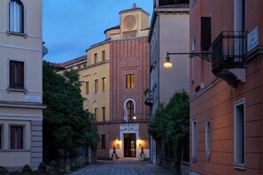 Une rue étroite bordée de bâtiments des deux côtés, dont un imposant bâtiment en briques rouges surmonté d’une tour horlogère et portant une enseigne indiquant « Calle Della Carmine ».