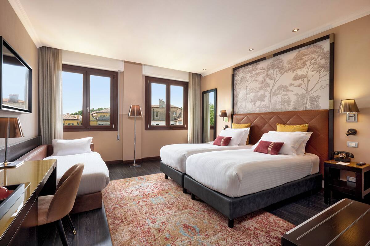 Une chambre d’hôtel avec deux lits, un bureau, une chaise, une grande fenêtre et un tableau au mur.