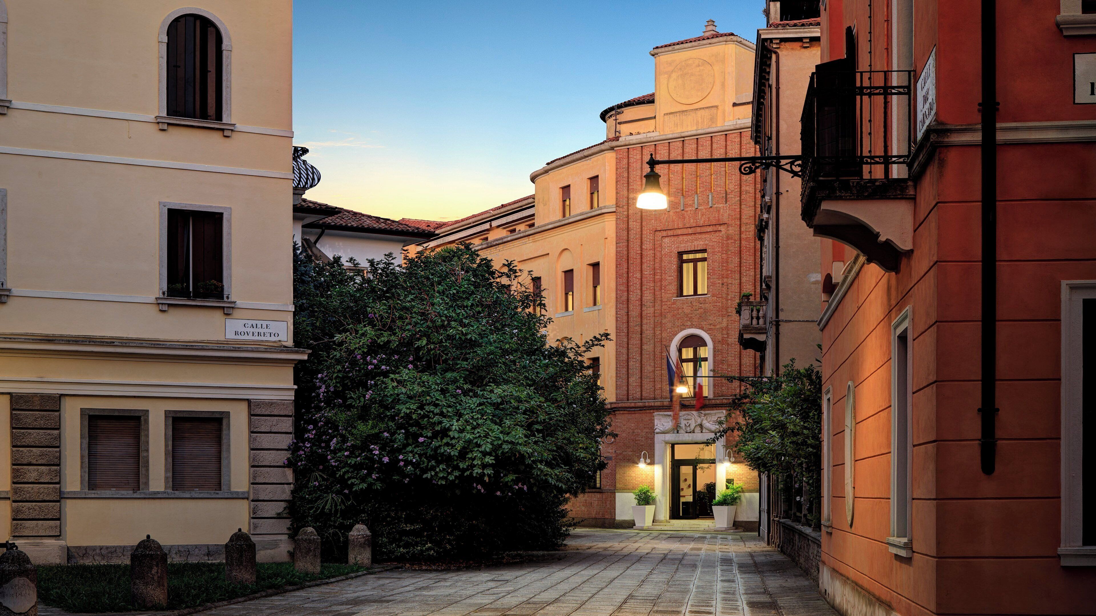 Un vicolo stretto fiancheggiato da edifici, con un albero al centro e un cartello che riporta la scritta 'Calle Rovereto'.